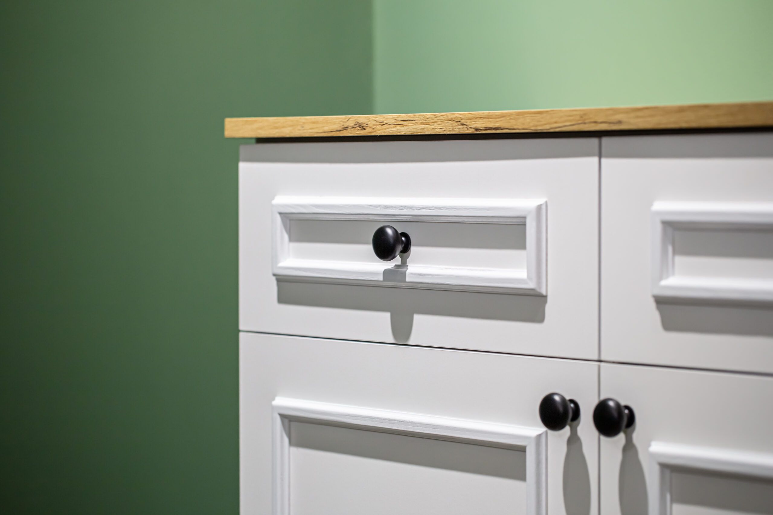 Close-up of a white kitchen cabinet with black handles against a green wall background.